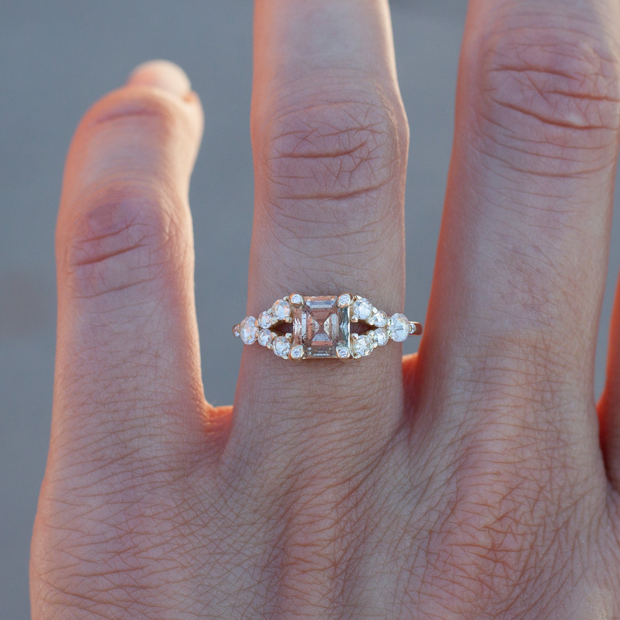 Unique Emerald cut diamond engagement ring, featuring a salt and pepper diamond, prong set, with small diamonds nested in the prongs of the setting. The center stone sits on a split band, featuring round rose cut and brilliant cut diamonds. Made in 14k or 18k gold.